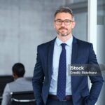 Portrait of handsome mid adult business man standing in modern office. Successful mature entrepreneur in formal clothing looking at camera with satisfaction. Confident man in suit with eyeglasses and beard standing with hands in pocket and looking at camera in office.